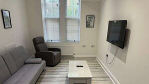 A living room with a grey sofa, a small armchair, a white coffee table, and a TV mounted on the wall. - Immaculate 1-bed Apartment in Walton on the Naze (Walton on the Naze)