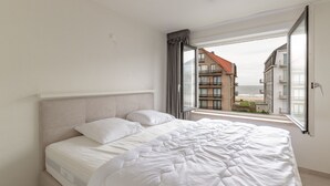 A bedroom with a large bed, a window with a view of buildings and the sea, and a ceiling light fixture. - Stylish Stay in De Haan (De Haan)