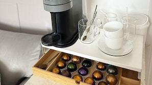 Una cafetera, un cartel con motivos de café y una caja de cápsulas de café sobre una mesa blanca.