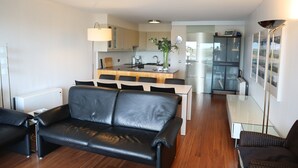 A modern living room with a black leather sofa, a dining table with chairs, and a kitchen area in the background. - Apartment in Nieuwpoort With sea View (Nieuwpoort)