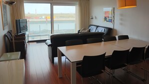 A hotel room with a large window, a sofa, a dining table with chairs, a TV, and a lamp. - Apartment in Nieuwpoort With sea View (Nieuwpoort)