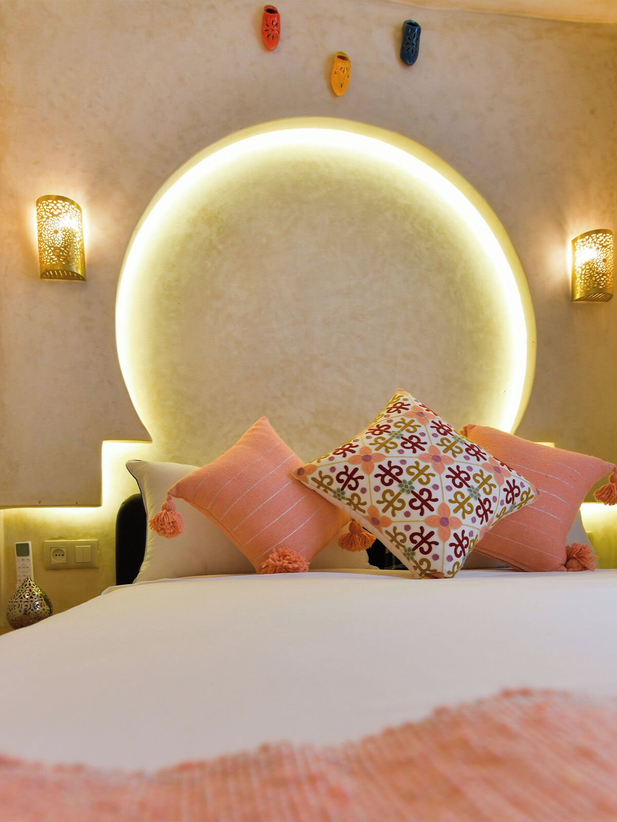 A hotel room with a large round mirror, a bed with multiple pillows, and wall-mounted sconces.