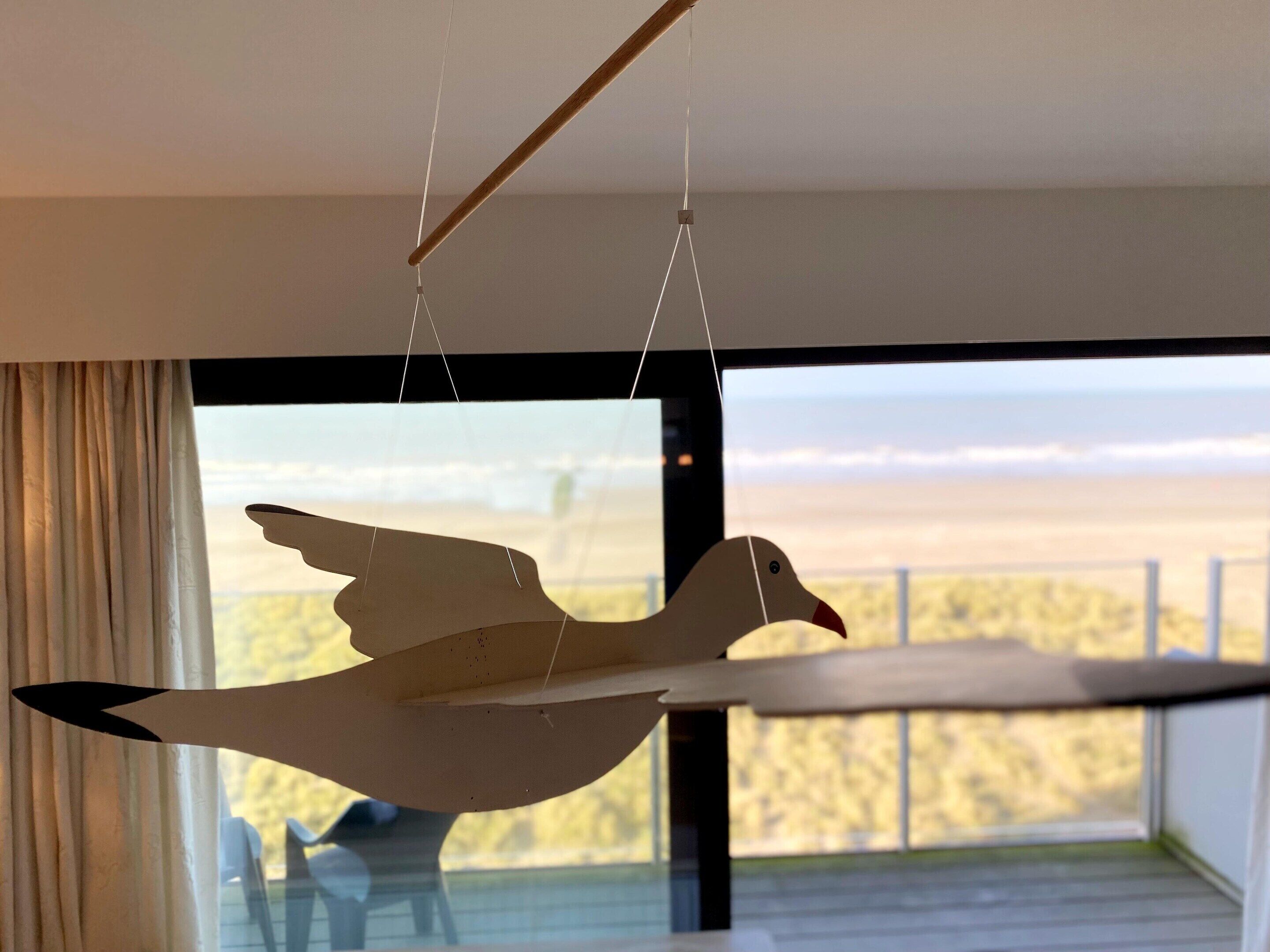 A room with a large window overlooking a beach, a hanging wooden bird sculpture, and a curtain partially drawn to the side.