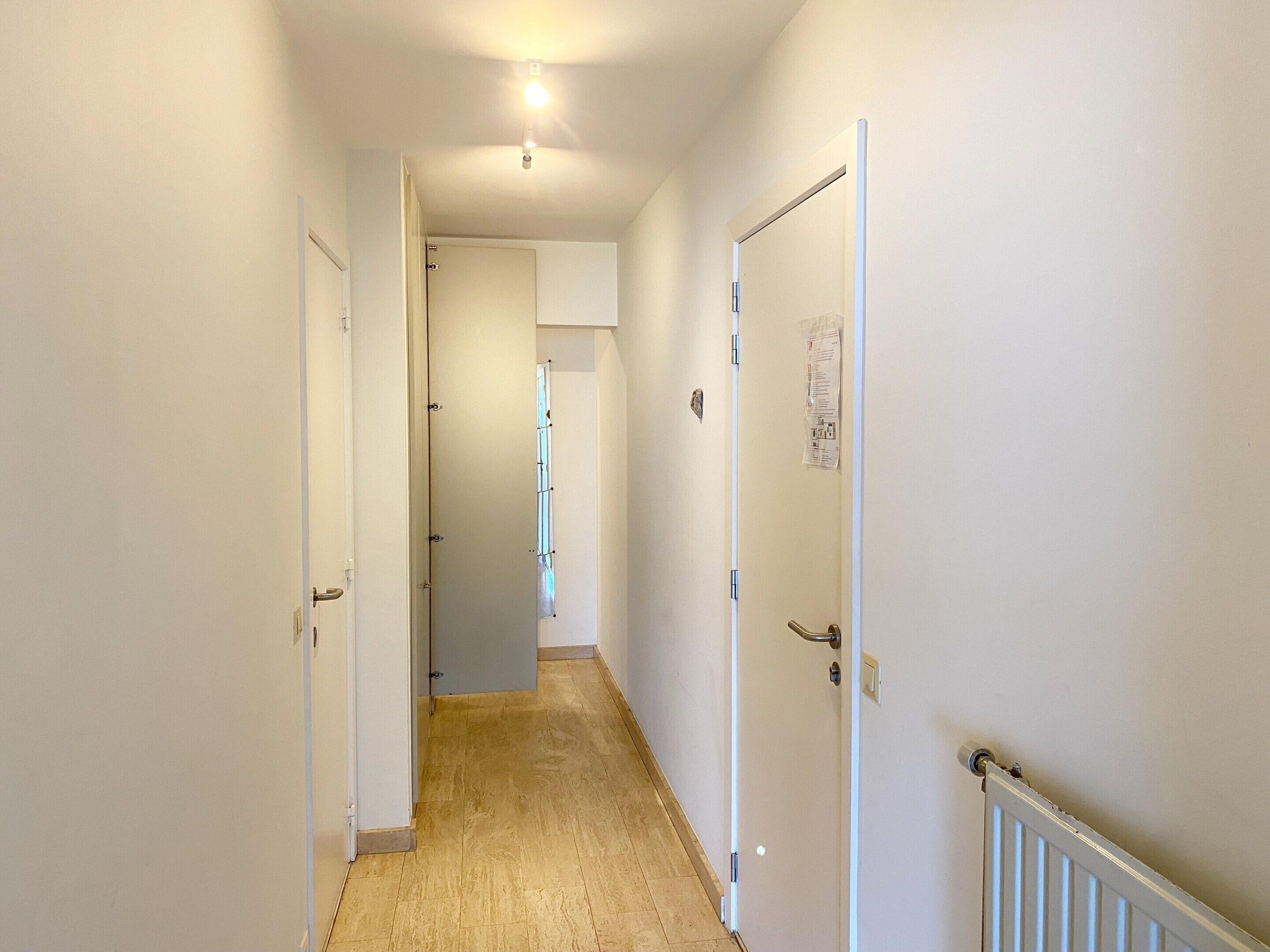 A narrow hallway with white walls and a wooden floor. There are doors on both sides, one of which has a bulletin board with papers attached.