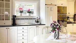 Een keuken met witte kastjes, een raam met bloemen, en een eettafel met een bloevas.