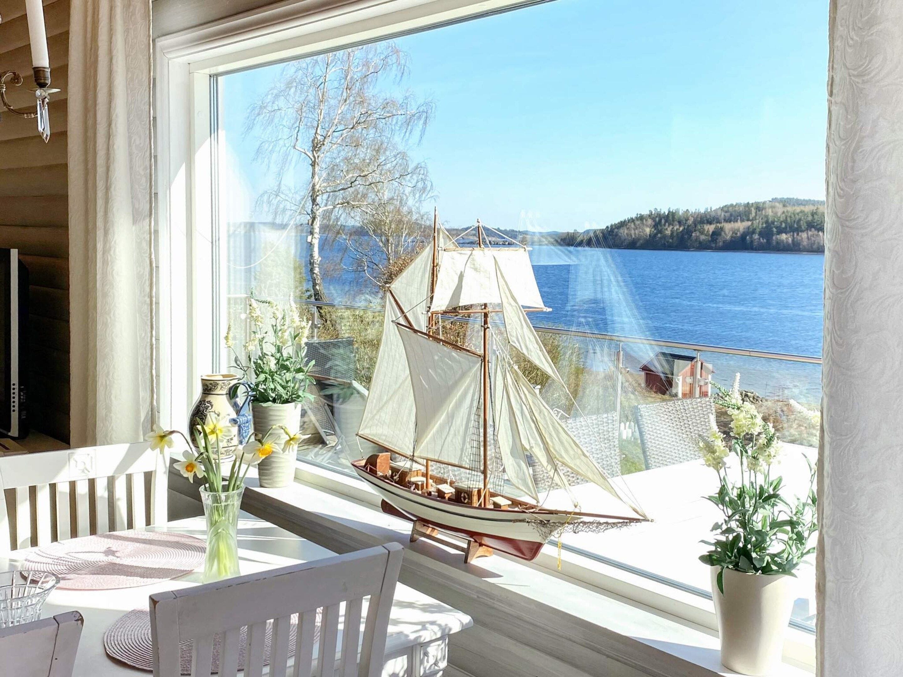 A room with a view of a lake, a model sailboat, and potted plants on a windowsill.