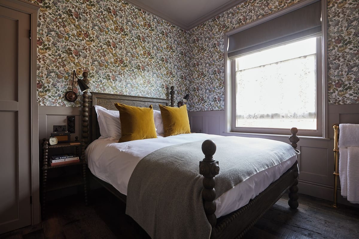 A bedroom with a floral wallpaper, a wooden bed with a headboard, two pillows, a bedside table with a radio, and a window with a view.