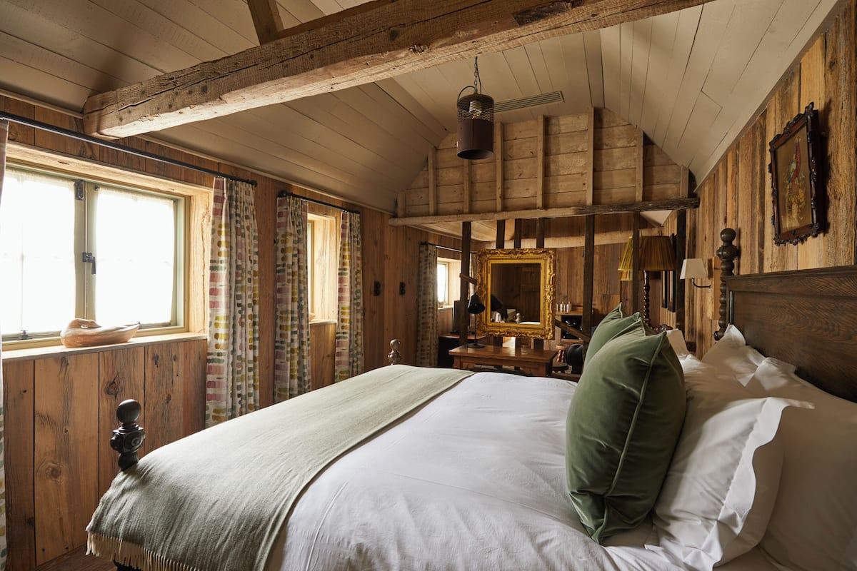 A wooden cabin bedroom with a large bed, a window with curtains, a wooden dresser, and a mirror on the wall.
