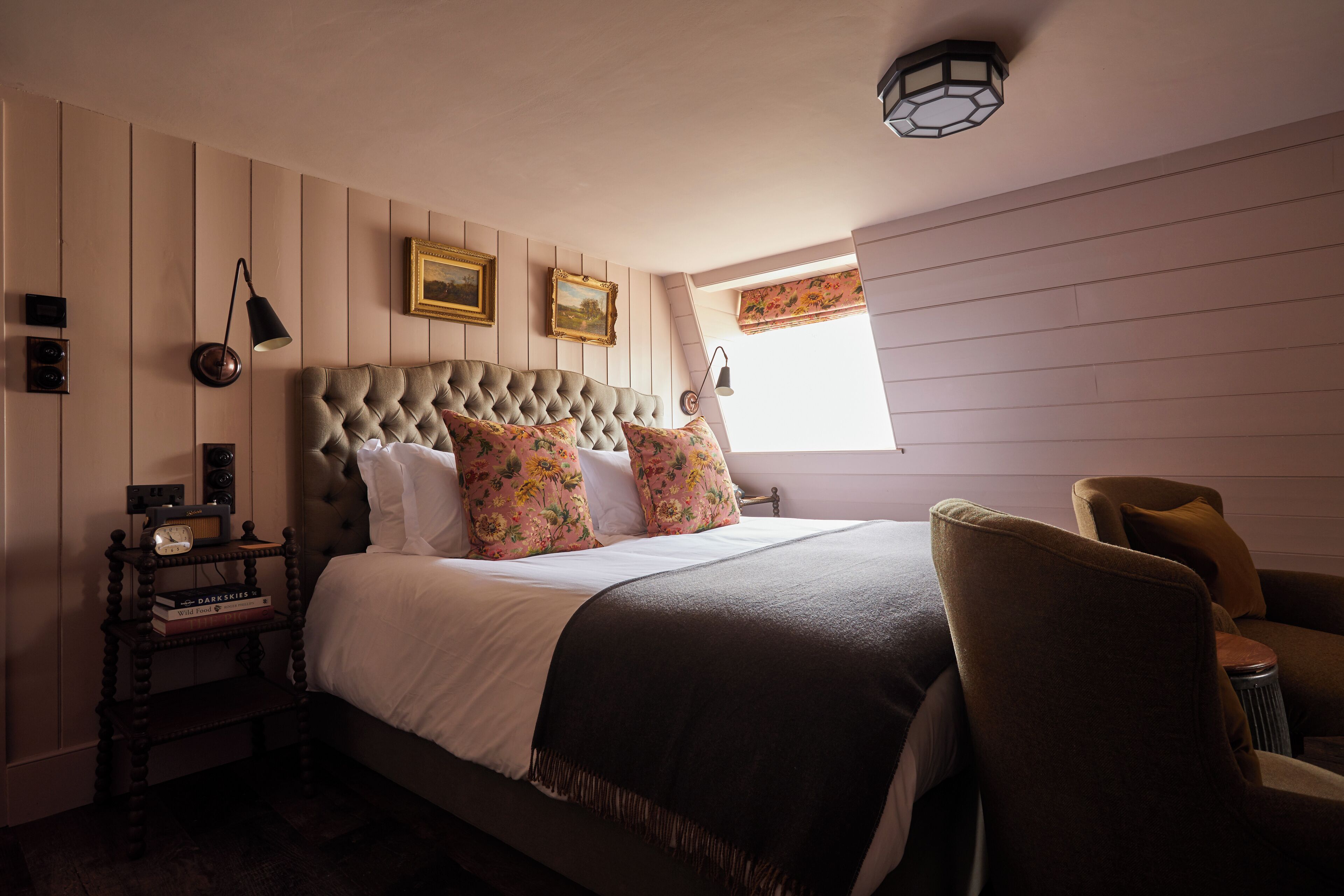 A bedroom with a tufted headboard, floral pillows, a bedside table with books, and a nightstand.