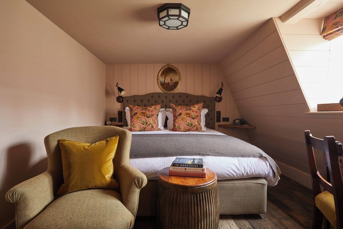 A cozy bedroom with a tufted headboard, floral pillows, a bedside table with books, and a sloped ceiling.