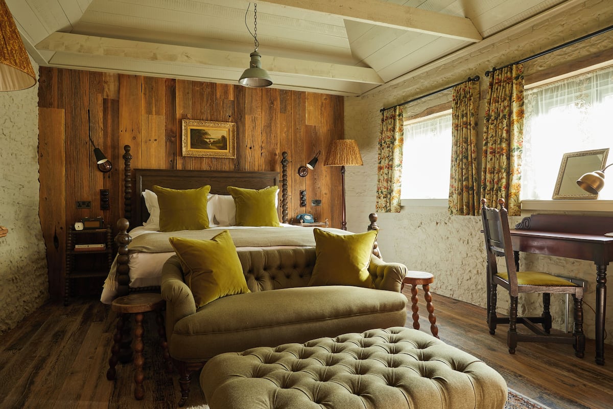 A bedroom with a wooden bed, green pillows, a tufted ottoman, a wooden nightstand, a desk, and a chair.