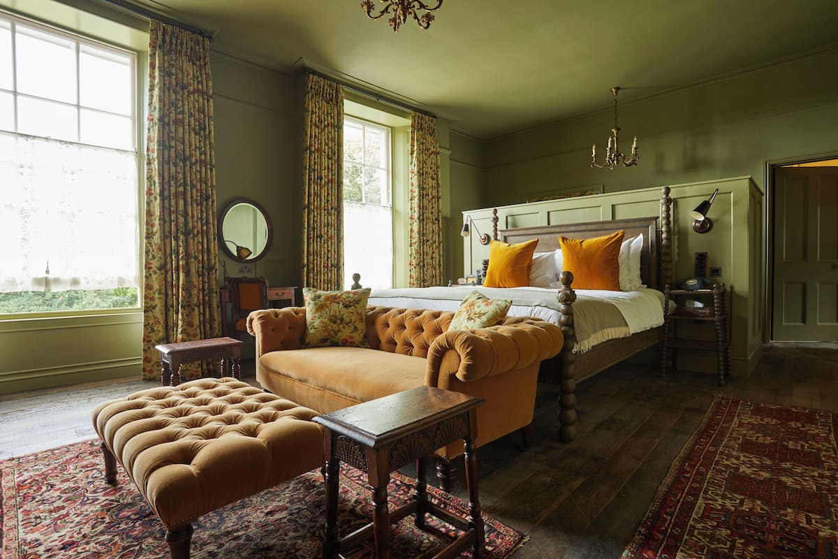 A bedroom with a large bed, a sitting area with a sofa and ottoman, a wooden nightstand, and a large window with curtains.