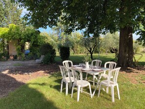 Un área de comedor al aire libre con una mesa dispuesta para cuatro personas, rodeada de sillas de plástico blanco, bajo un árbol grande.