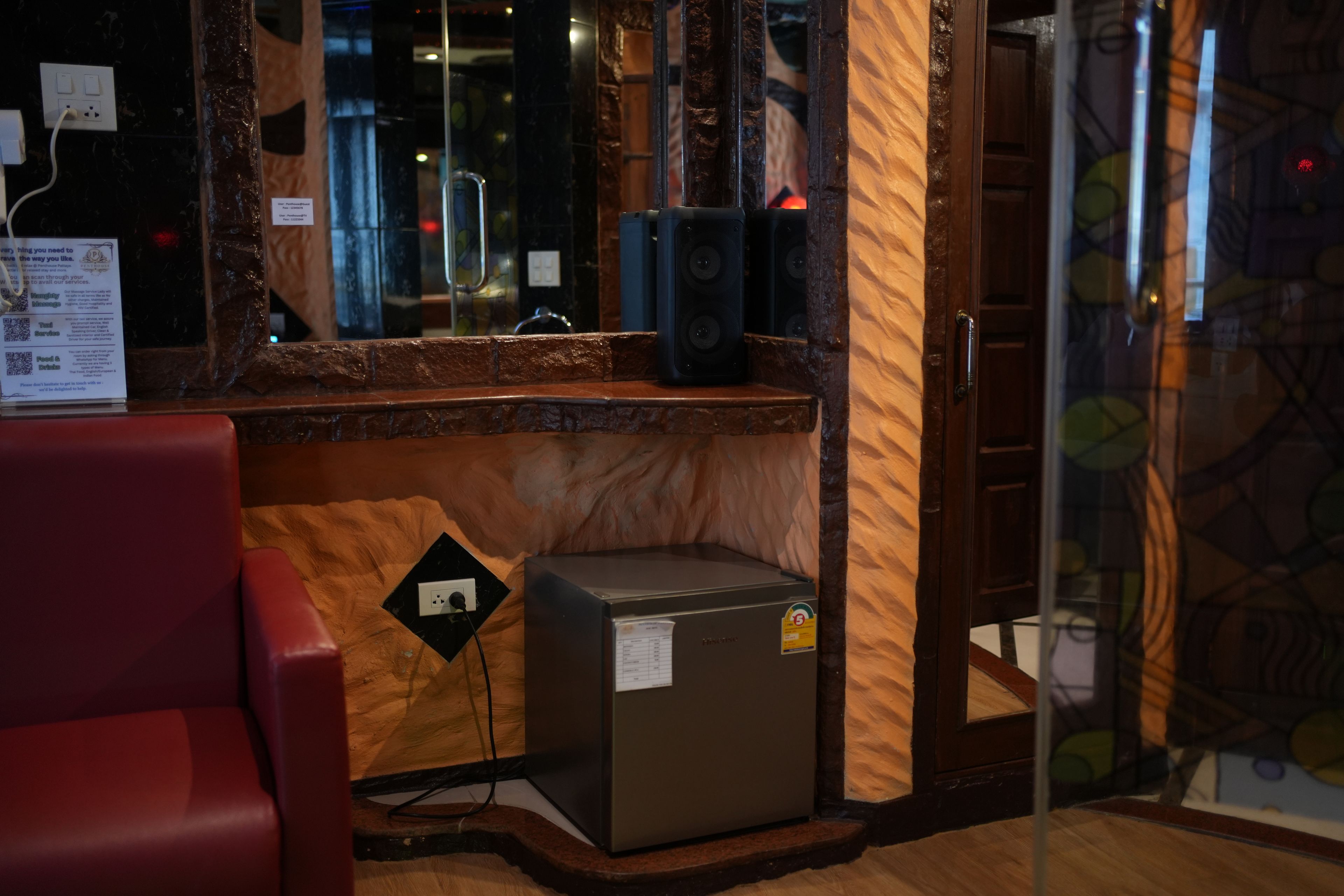 A room with a wooden counter, a small refrigerator, a red chair, and a mirror reflecting the room.