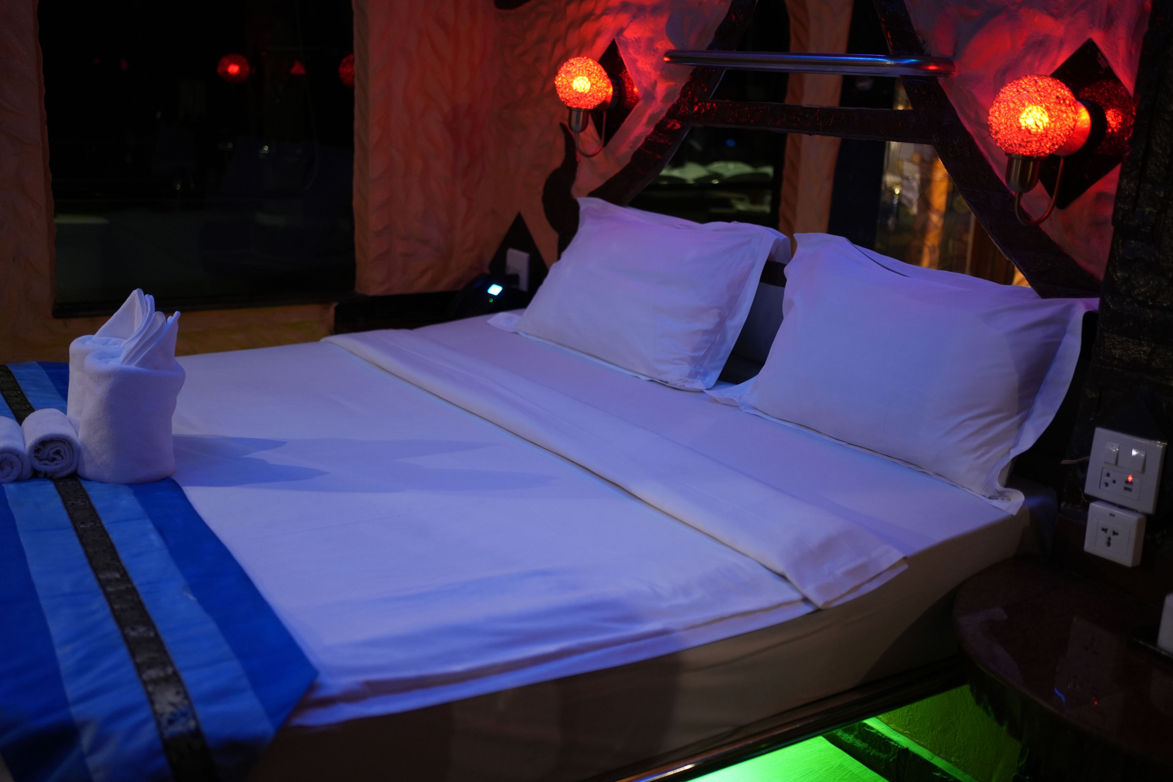 A hotel room with a bed, pillows, a bedside table with a lamp, and a window.