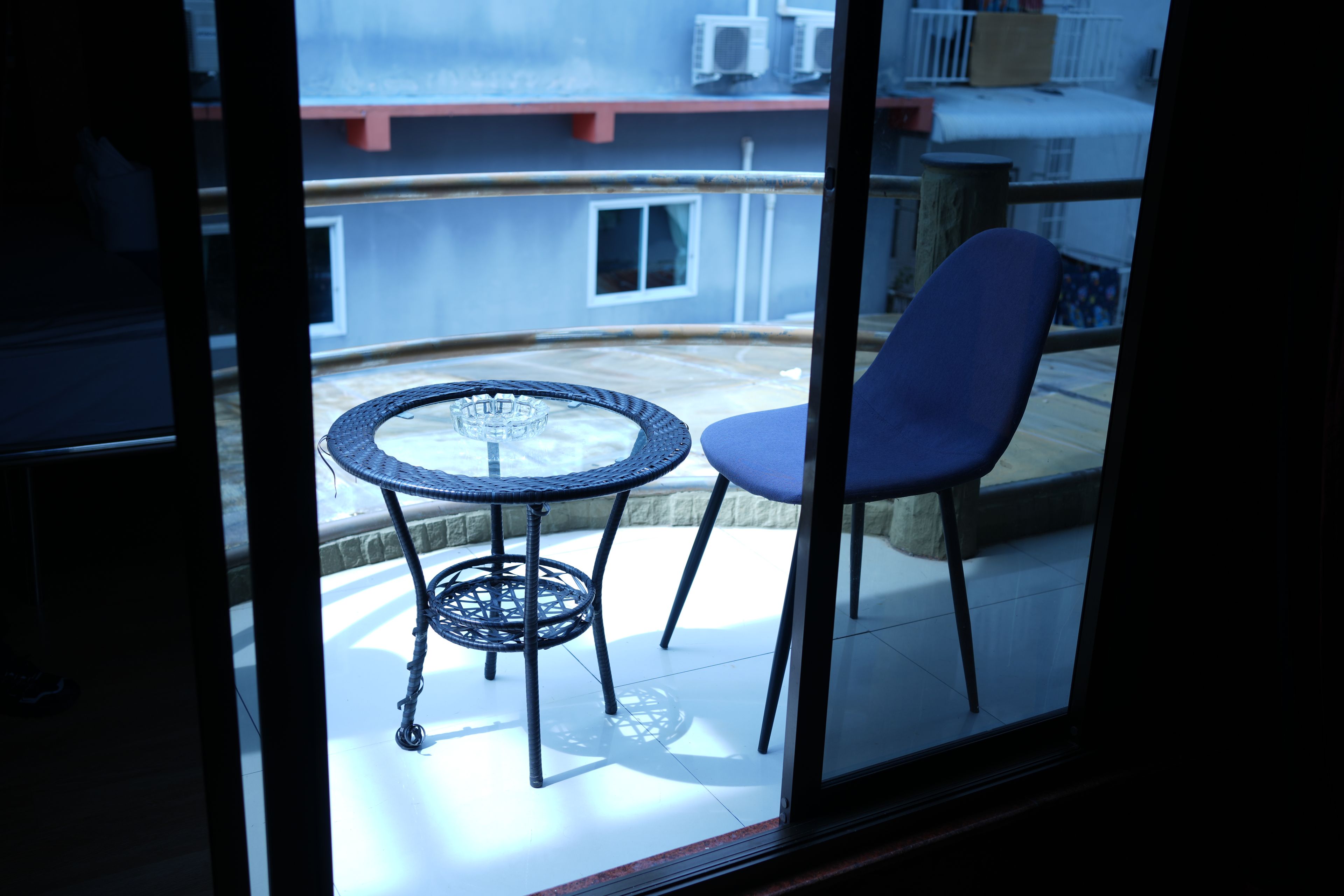 Un balcon avec une table ronde et deux chaises, une vue sur un immeuble équipé de climatiseurs et une rambarde.