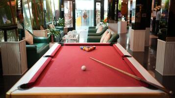 A pool table with balls and a cue stick, surrounded by green sofas and a glass partition.