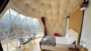 Een slaapkamer met een groot raam, een bed, een nachtkastje en een kleine tafel met een stoel.
