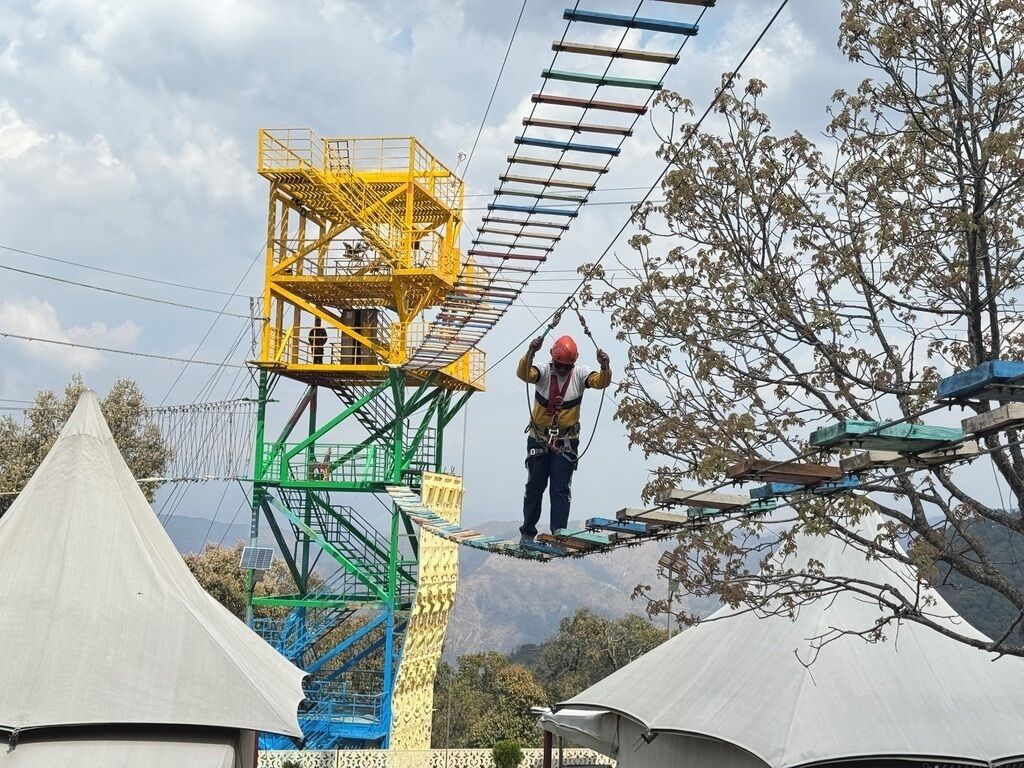 A person on a rope bridge with a yellow and green metal structure in the background.