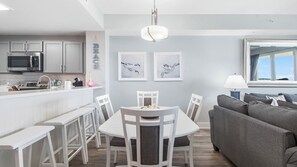 A modern kitchen with stainless steel appliances, a dining area with white chairs and a table, and a grey sofa in a bright living room with artwork on the walls. - Calypso 3-1606 Spectacular condo! Affordable rates! Inquire for details! (Panama City Beach)