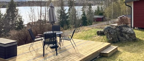 Um deck de madeira com móveis para área externa, vista para um lago e um prédio vermelho ao fundo.