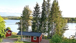 Une cabane rouge avec un porche abrité, entourée d’arbres et bordée par un lac.