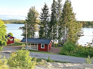 Punainen mökki, jossa on katettu kuisti, ja sen ympärillä on puita ja järvi.