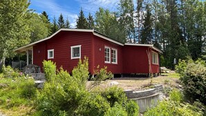 Une maison en bois rouge, dotée d’un porche, entourée d’une végétation luxuriante et d’arbres.