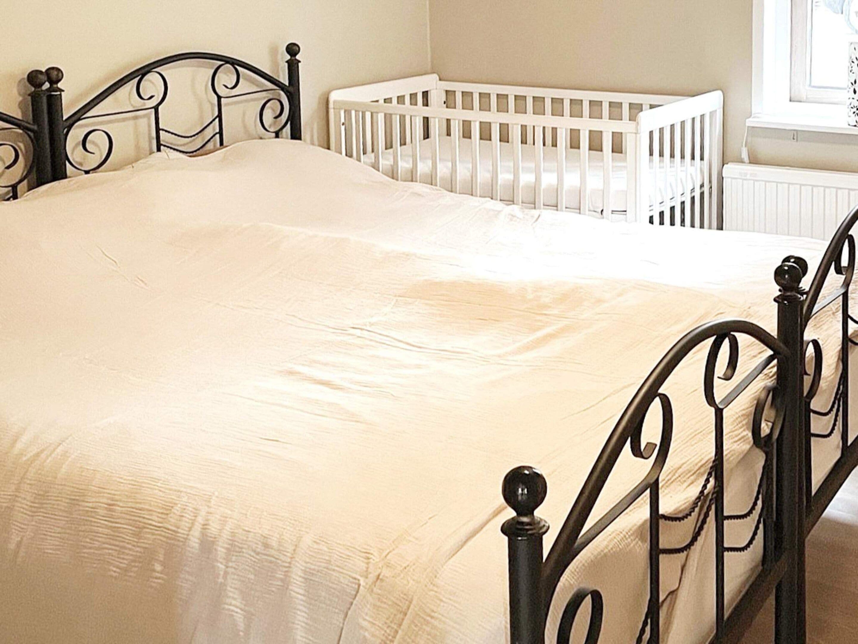 Una cama con colchón blanco y estructura metálica negra en una habitación con una cuna blanca y un ventanal.