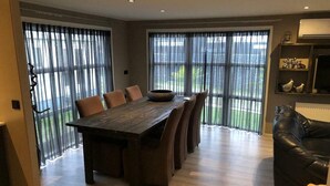 A spacious dining area with a long wooden table and chairs, large windows with sheer curtains, a black leather sofa, and a radiator on the wall. - Holiday Home on the Recreational Lake (Lathum)