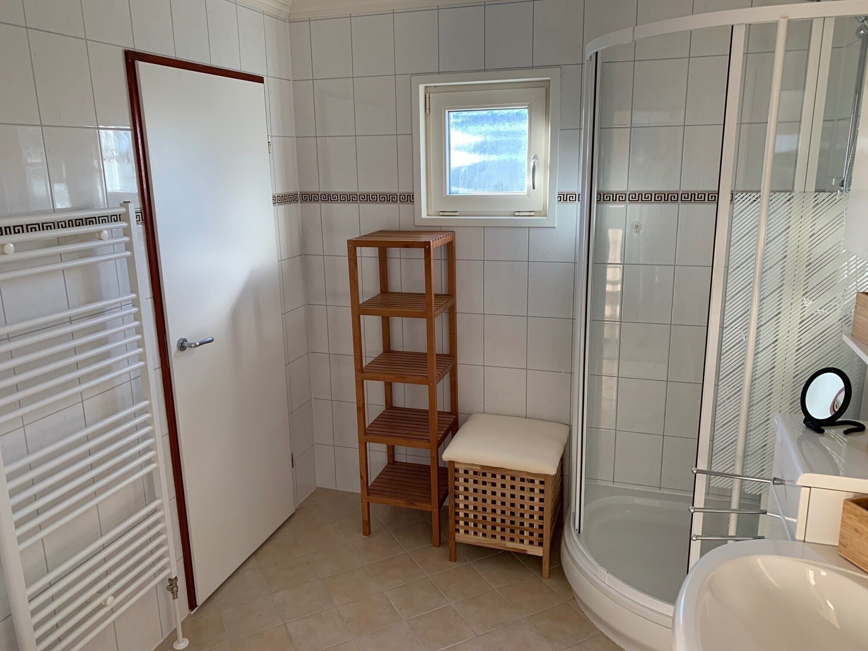 A bathroom with a shower, a toilet, a sink, a wooden shelf, and a small window.