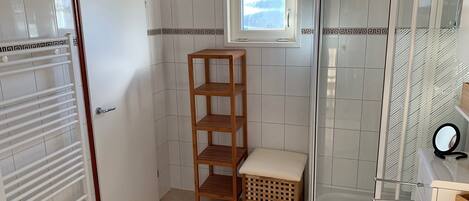 A bathroom with a shower, a toilet, a sink, a wooden shelf, and a small window.