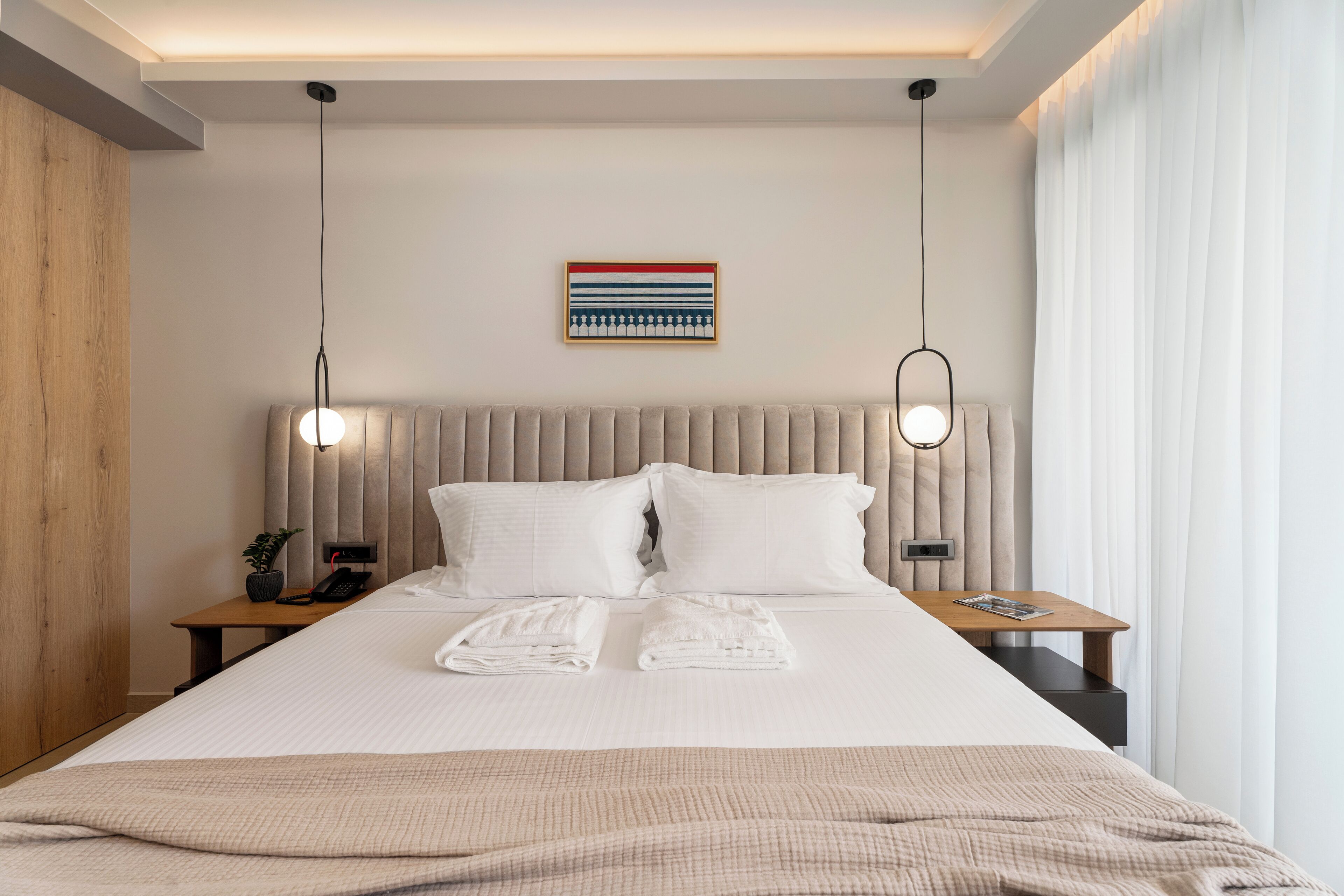 Una cama bien hecha con dos almohadas y dos toallas dobladas, una mesita de noche con una lámpara, una planta pequeña y un cuadro en la pared.