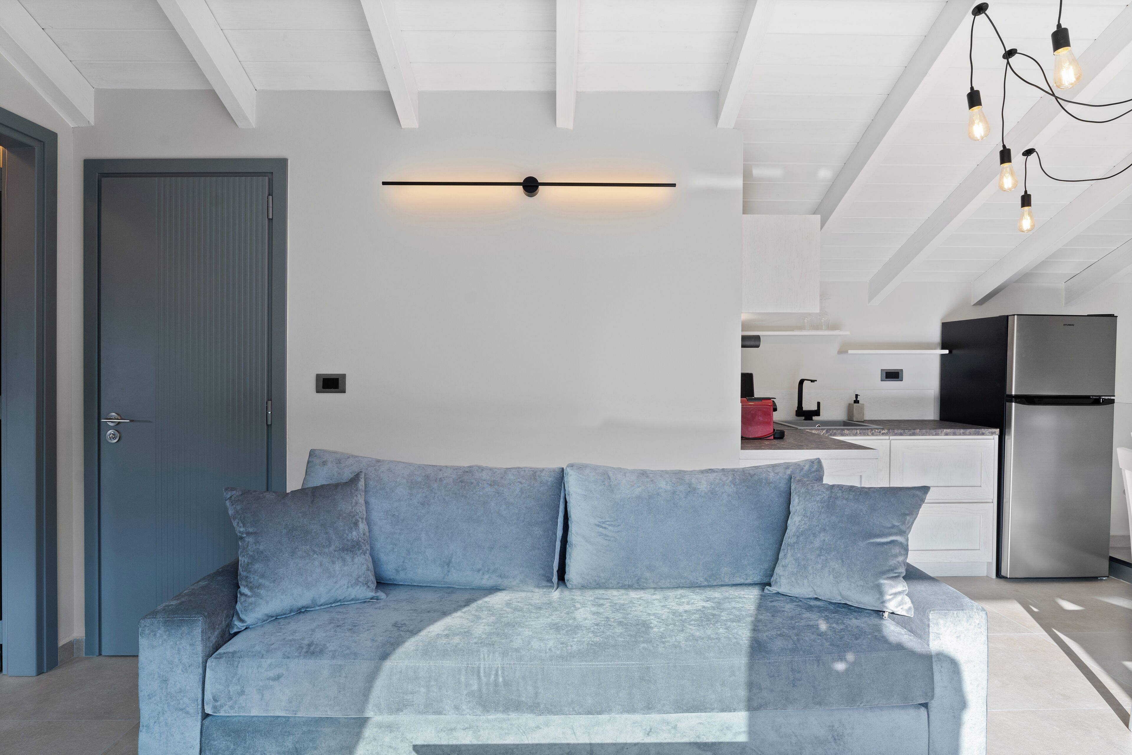 Una sala de estar moderna con un sofá gris, una lámpara de techo y una cocina al fondo.