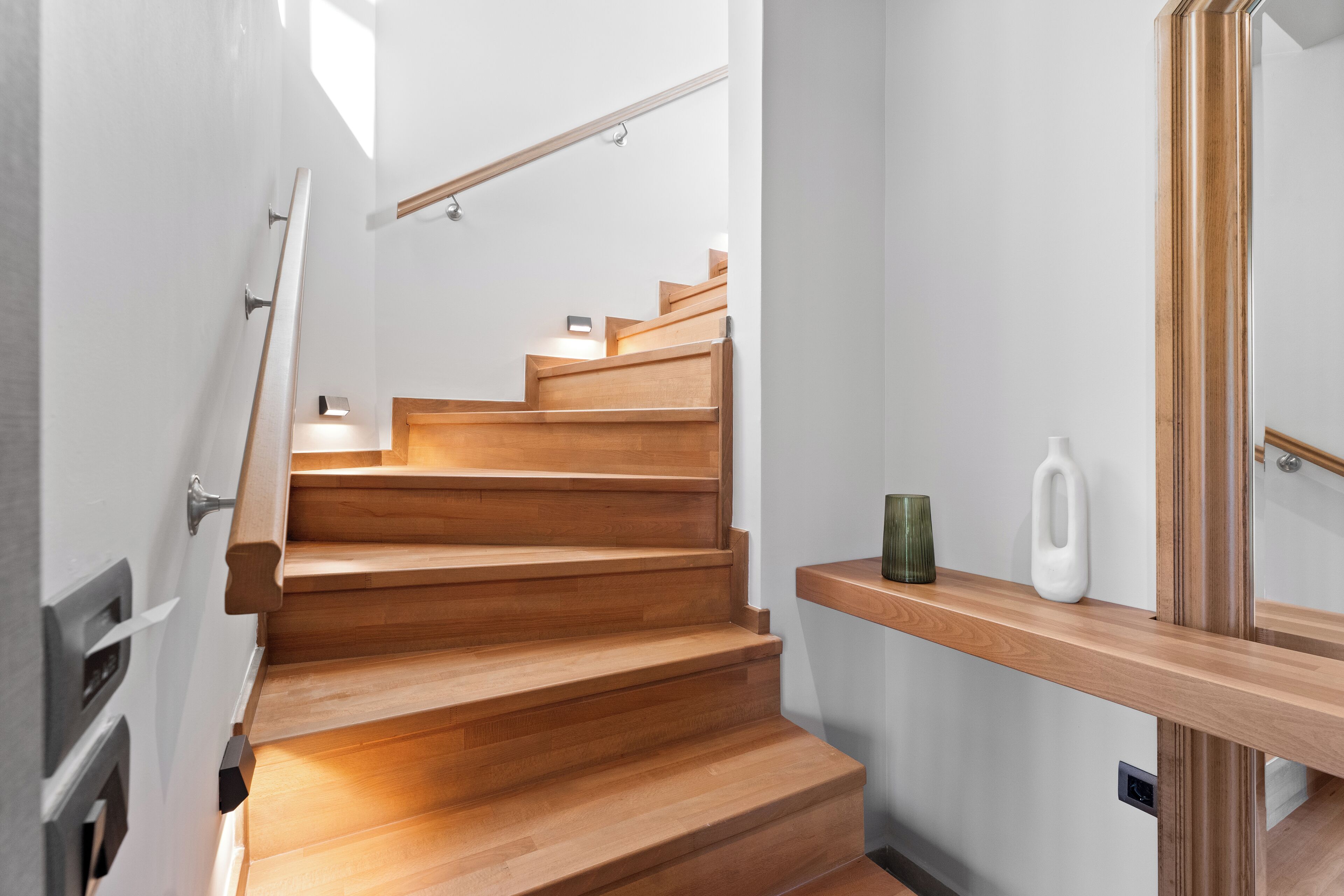 A wooden staircase with a handrail, a small shelf with a vase and a bottle, and a mirror on the wall.