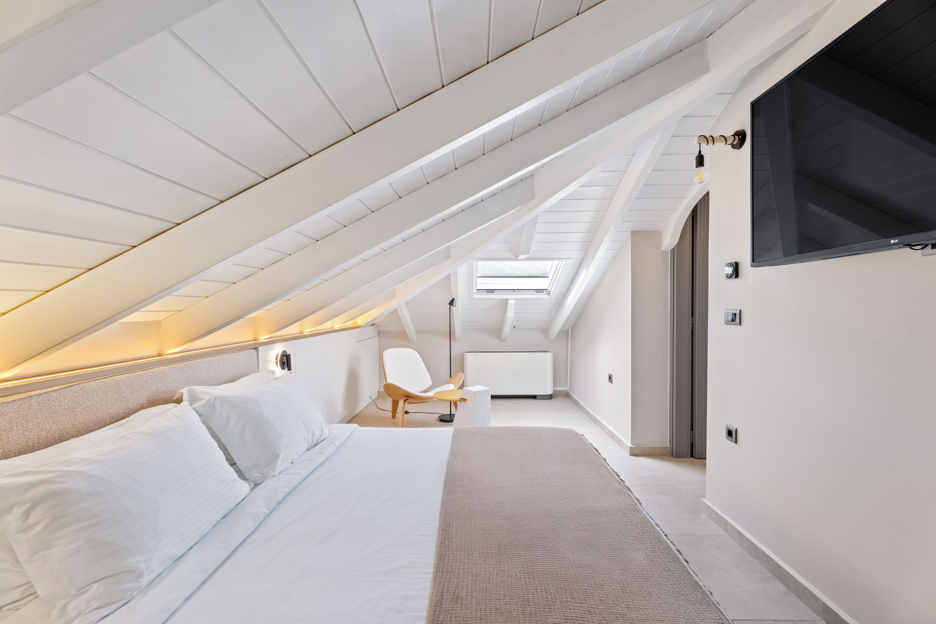 A modern bedroom with a sloped ceiling, a bed, a chair, and a TV mounted on the wall.