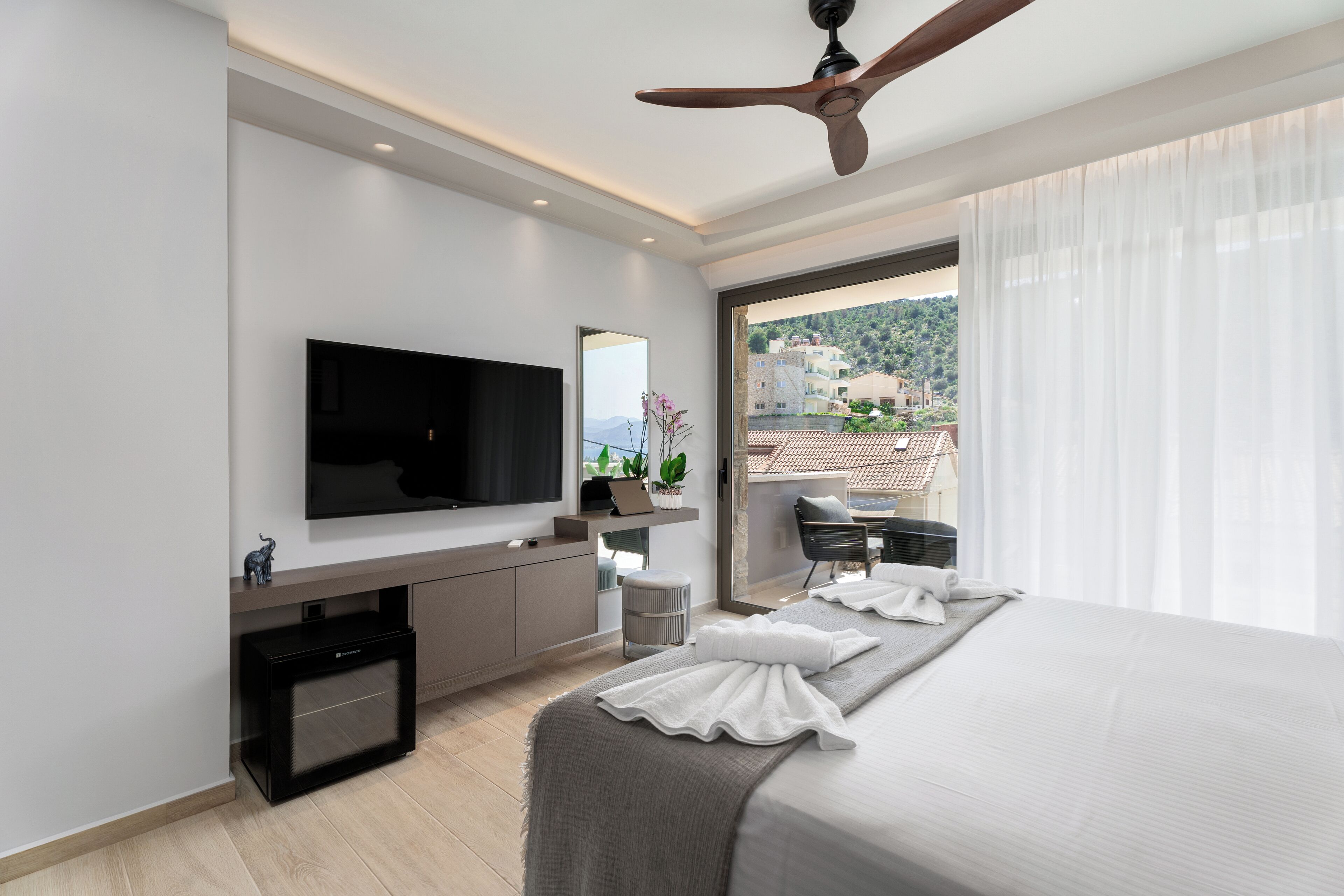 A modern hotel room with a flat-screen TV, a ceiling fan, a balcony with a view of the city, and a neatly made bed.