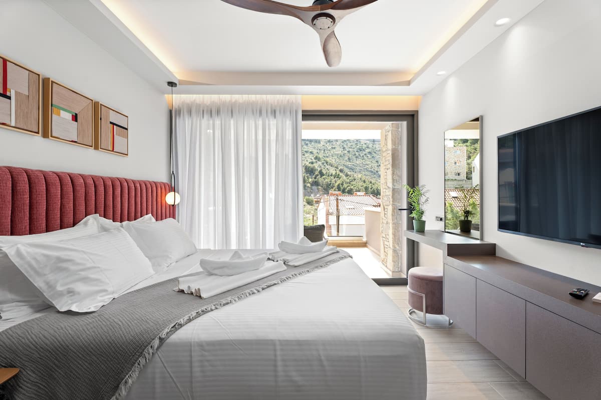 A modern bedroom with a large bed, a TV, and a balcony with a view of the countryside.