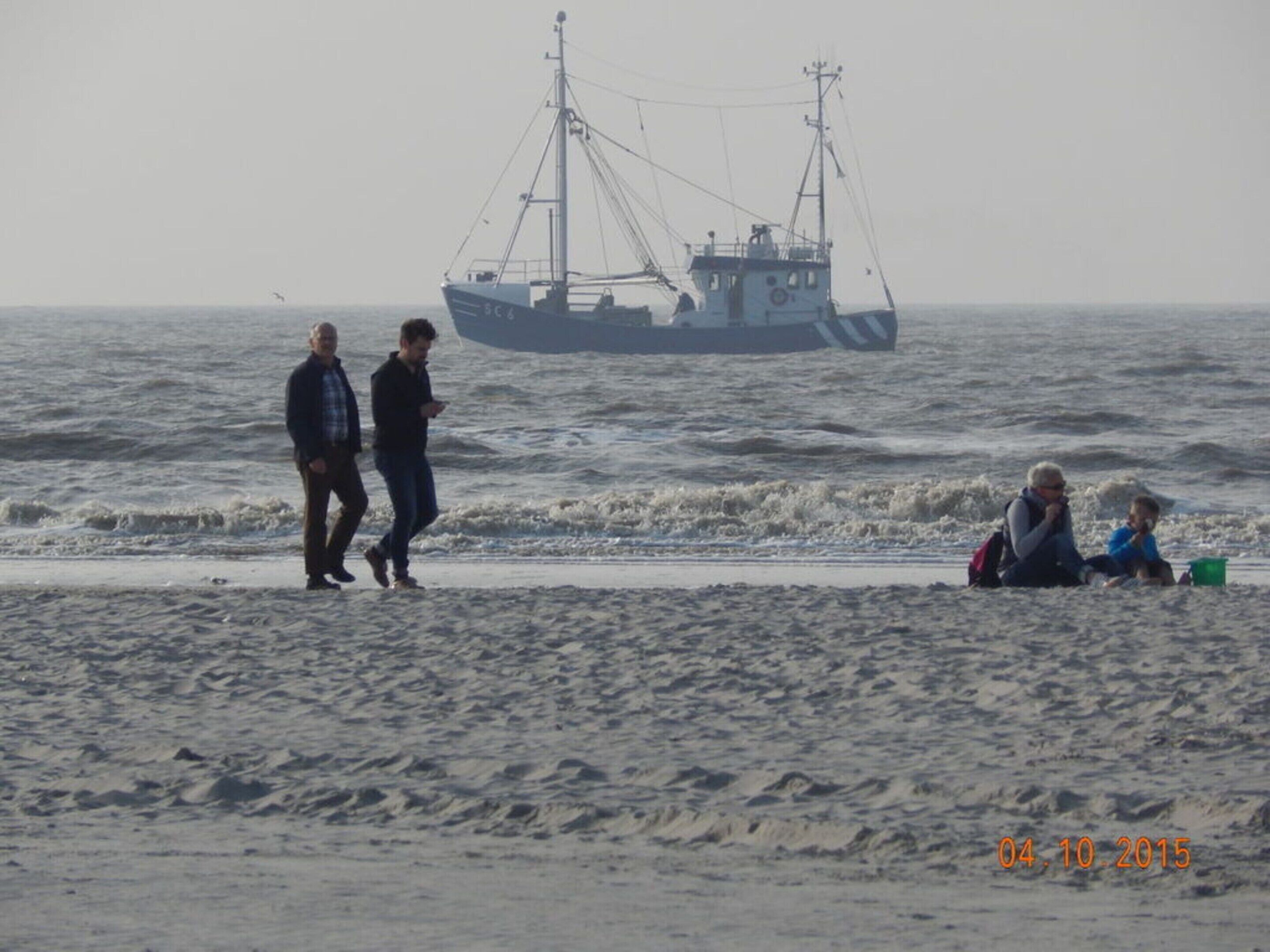 Une plage avec un bateau de pêche en arrière-plan, et des gens qui se promènent et s'assoient sur le rivage.
