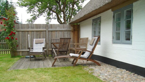 Een terras met drie houten stoelen en een tafel, een wit gebouw met een zwarte plint, en een houten hek op de achtergrond.