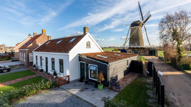 En traditionel vindmølle og et hvidt hus med tegltag, omgivet af en grusvej og grøntområder.