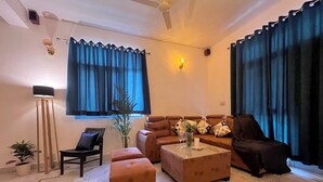 A living room with a sofa, ottoman, floor lamp, and a potted plant. - Rangoon by Grehstay 2BHK Independent House near Expo Mart (Greater Noida)