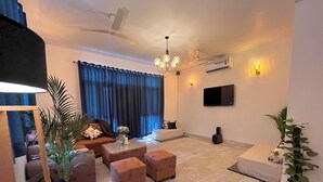 A modern living room with a sofa set, ottomans, a coffee table, and a TV mounted on the wall. - Rangoon by Grehstay 2BHK Independent House near Expo Mart (Greater Noida)