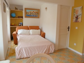 A bedroom with a bed, a chair, a shelf with decorative items, and a door.