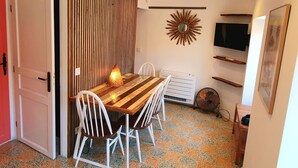 Un área de comedor con una mesa de madera y sillas blancas, un televisor montado en la pared, un espejo decorativo y un ventilador en el suelo.