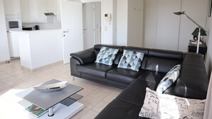 Ein modernes Wohnzimmer mit einer schwarzen Ledersofa, einem Couchtisch und einem durchgehenden Gang sichtbaren Küchenbereich.