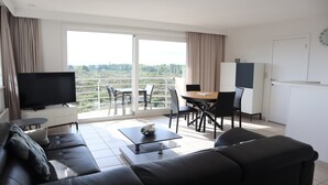 Ein modernes Wohnzimmer mit Sofa, Couchtisch, Fernseher, einem Balkon mit Essbereich und Blick ins Grüne.