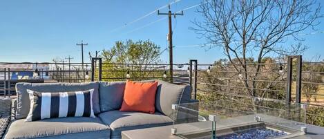 A modern outdoor seating area with a gray sofa and a glass table, overlooking a landscape with trees and power lines.