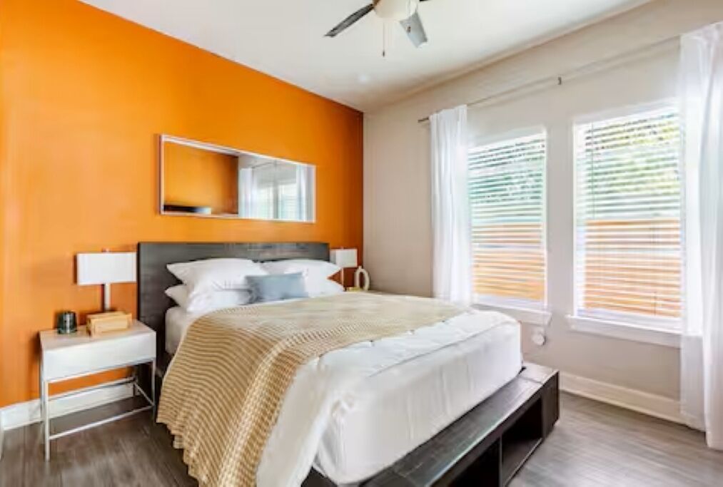 A bedroom with an orange wall, a bed with white bedding, a bedside table with a lamp, and a window with blinds.