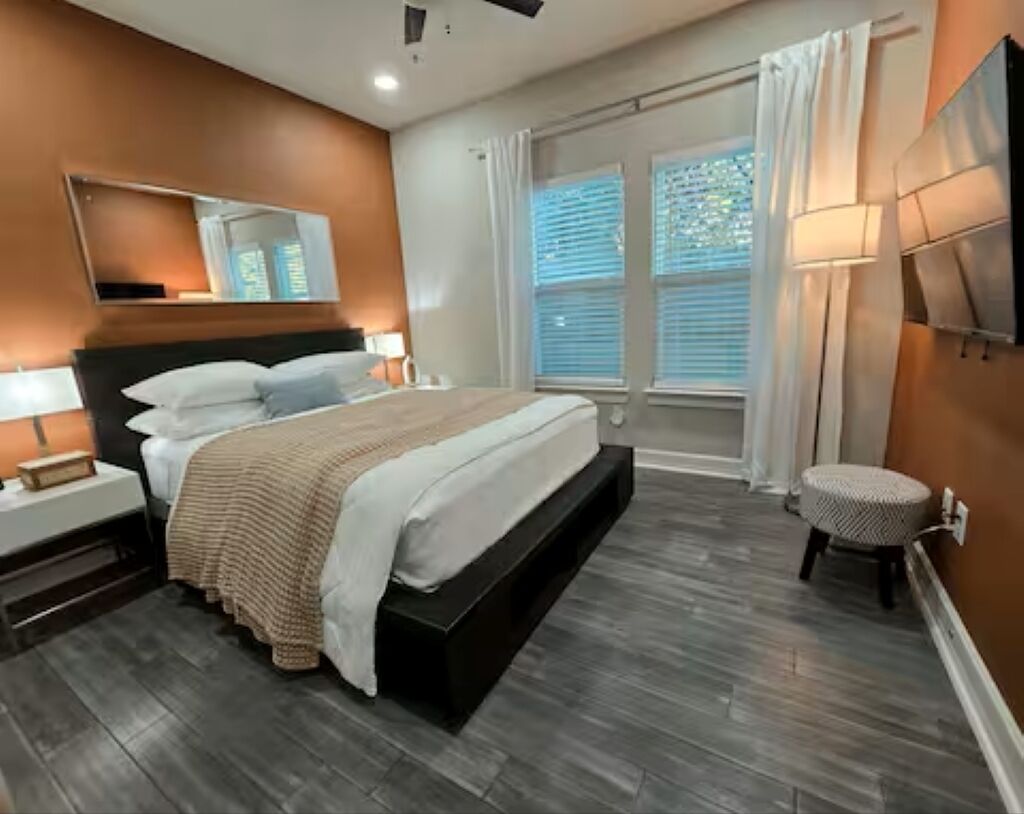 A modern bedroom with a large bed, a nightstand, a bedside table, a round stool, and a window with blinds.